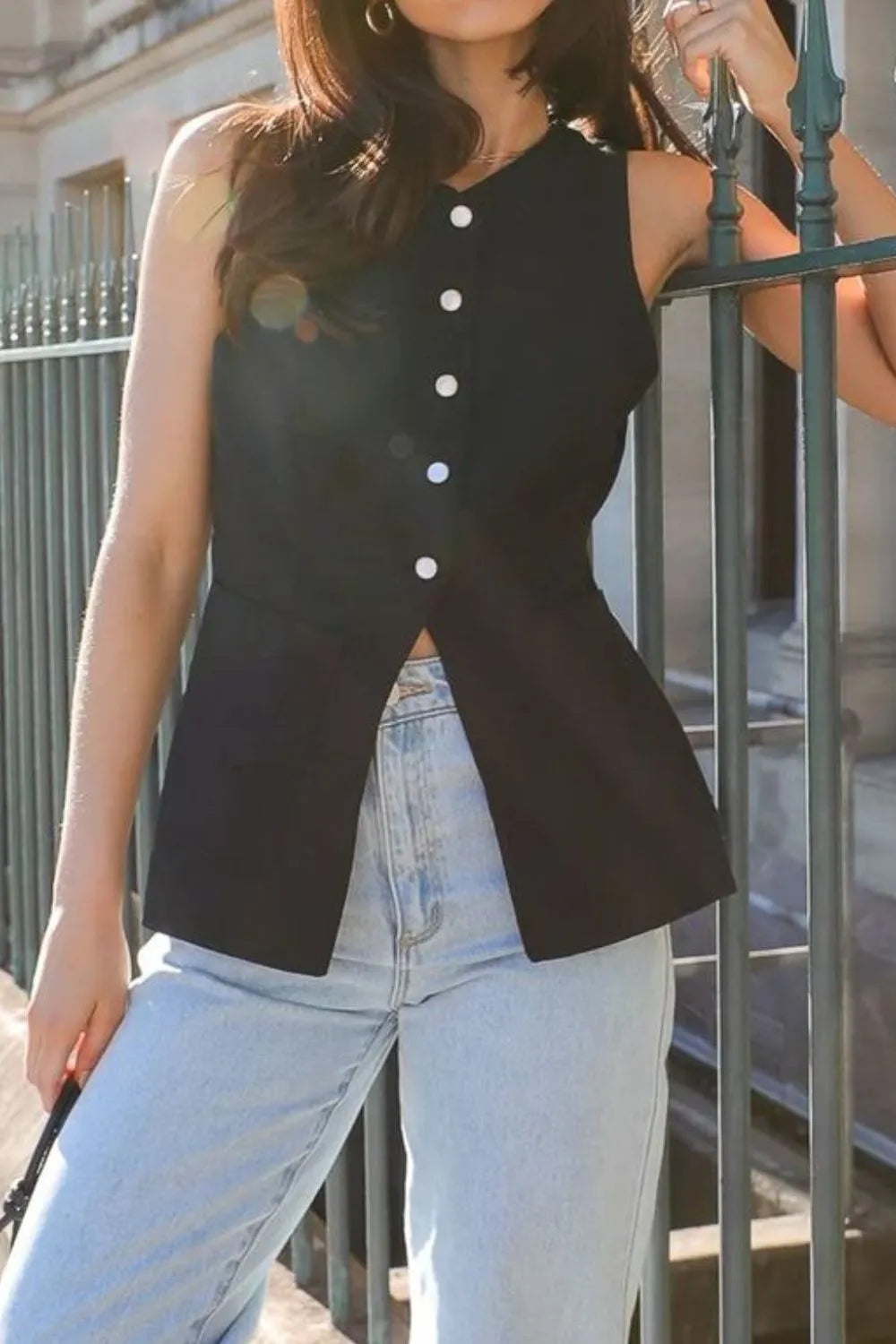 Person wearing a black sleeveless button-up top and light blue jeans leaning against a fence.