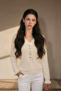 A woman with long dark wavy hair wearing a cream-colored knit henley sweater with button details and white pants, standing against a neutral beige wall.