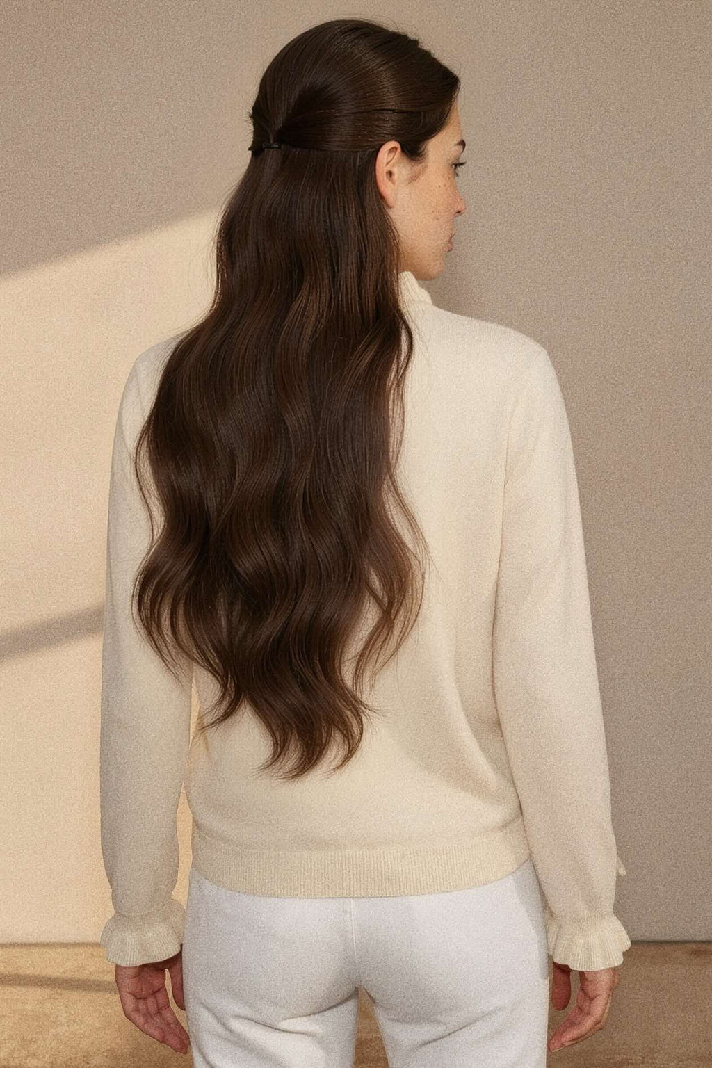 A woman shown from the back wearing a cream-colored knit sweater and white pants, with long, wavy brown hair. The background is a minimalist beige wall with soft shadows.