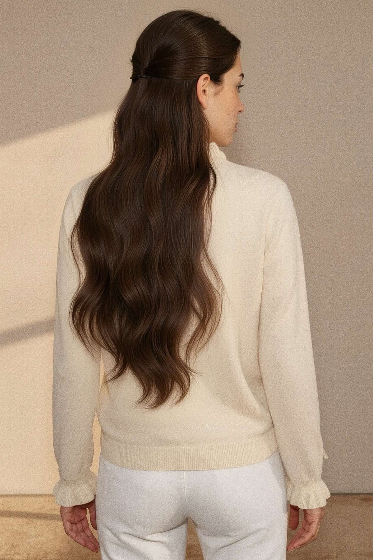 A woman shown from the back wearing a cream-colored knit sweater and white pants, with long, wavy brown hair. The background is a minimalist beige wall with soft shadows.