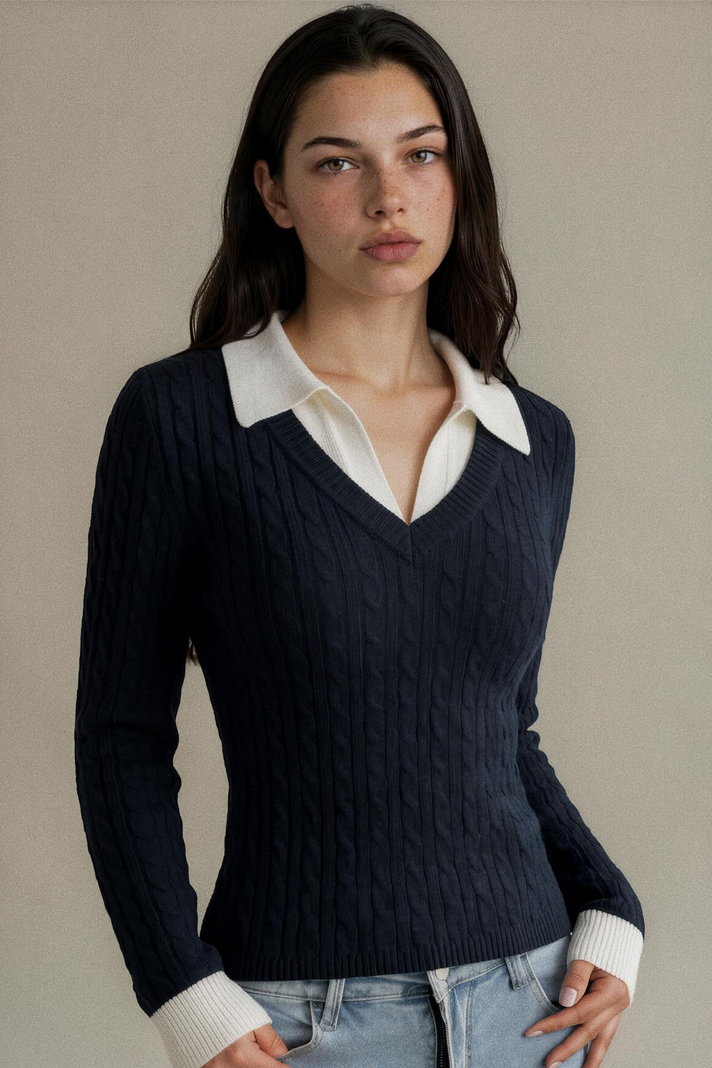 A woman wearing a navy blue cable-knit V-neck sweater with a contrasting white collar and white ribbed cuffs, paired with light blue jeans, set against a plain beige background.
