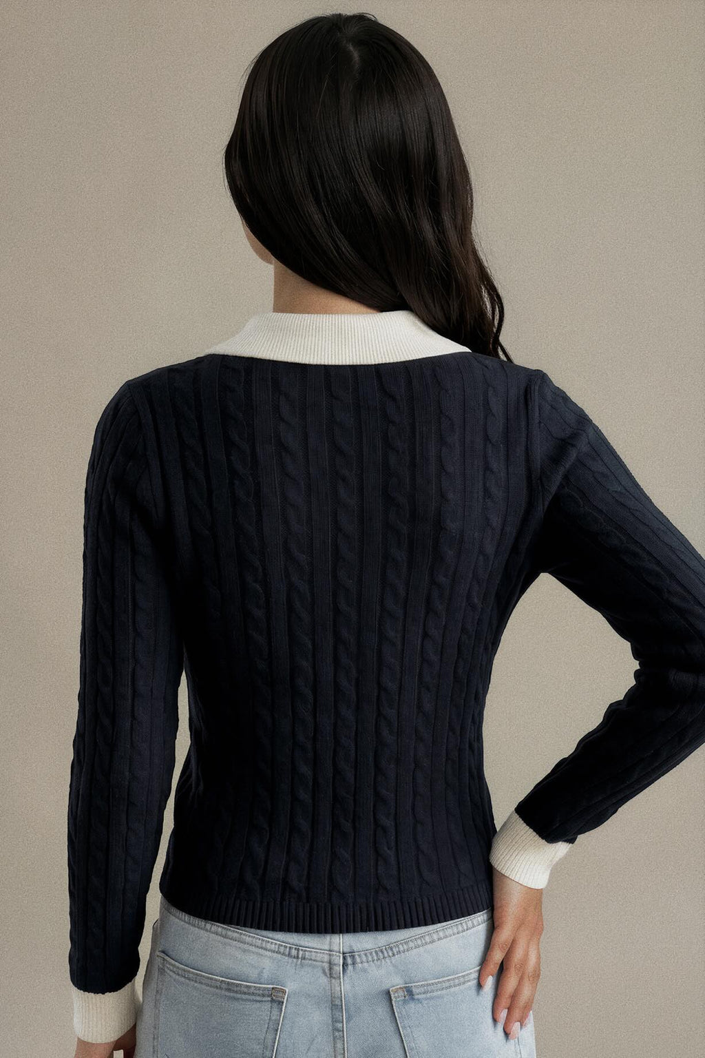 A woman shown from the back wearing a navy blue cable-knit sweater with contrasting cream ribbed collar and cuffs, paired with light-wash jeans against a neutral beige background.