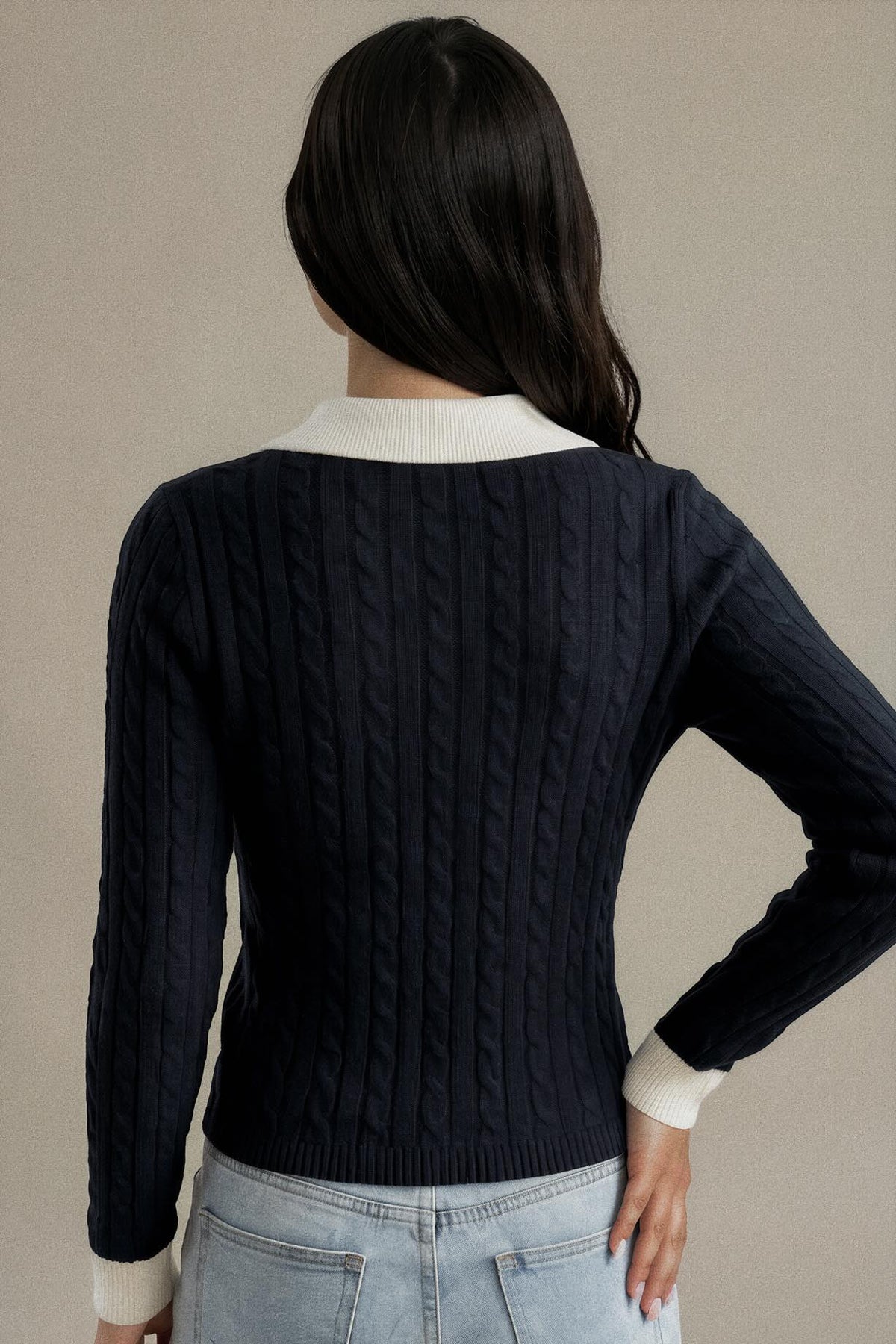 A woman shown from the back wearing a navy blue cable-knit sweater with contrasting cream ribbed collar and cuffs, paired with light-wash jeans against a neutral beige background.