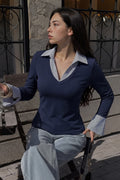 A woman wearing a navy blue long-sleeve V-neck sweater with light gray contrast collar and cuffs, paired with light gray trousers, sitting on a wooden chair outdoors.