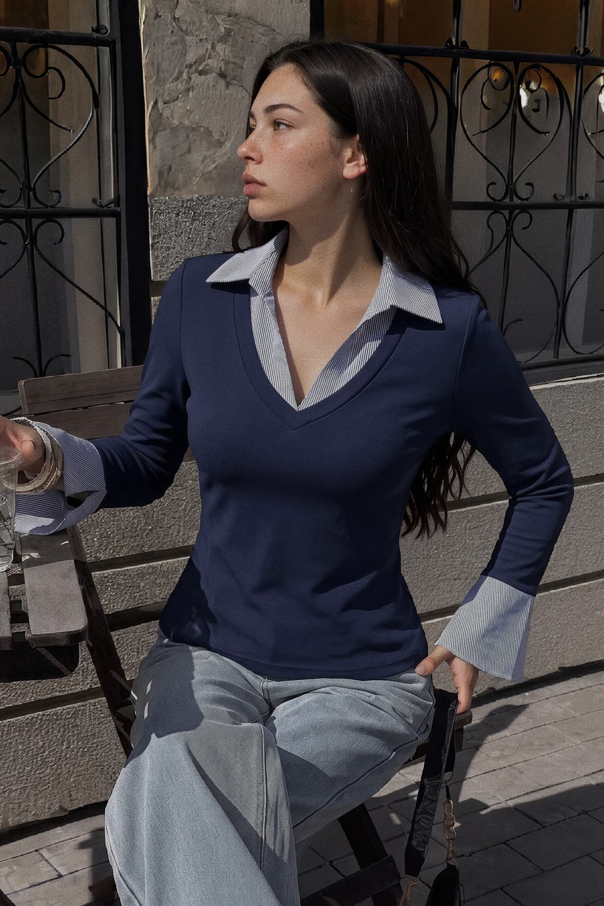 A woman wearing a navy blue long-sleeve V-neck sweater with light gray contrast collar and cuffs, paired with light gray trousers, sitting on a wooden chair outdoors.