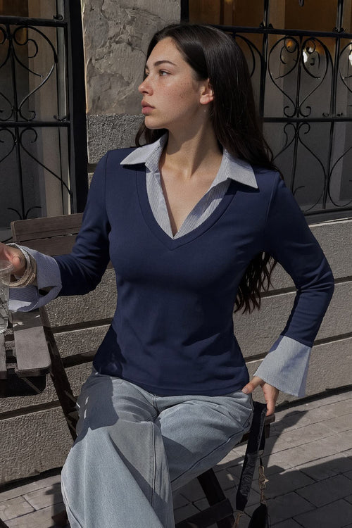 A woman wearing a navy blue long-sleeve V-neck sweater with light gray contrast collar and cuffs, paired with light gray trousers, sitting on a wooden chair outdoors.