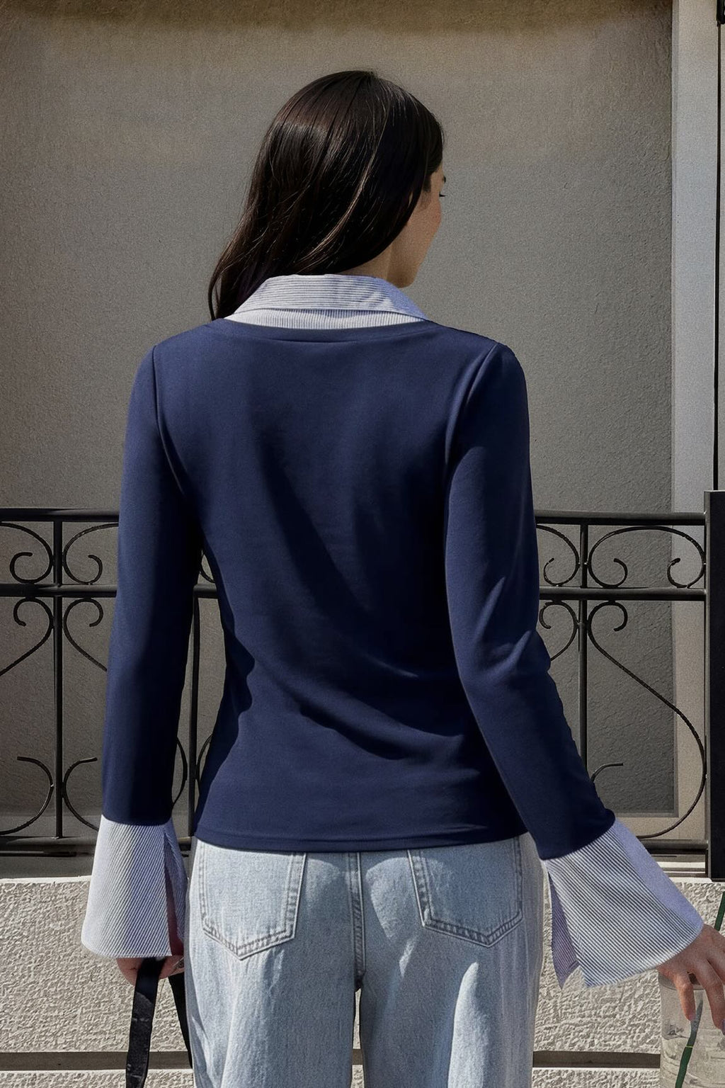 A woman viewed from behind wearing a navy long-sleeve top with contrasting light blue and white ruffled collar and cuffs, paired with light-wash jeans, standing outdoors in front of a beige wall and black wrought iron railing.