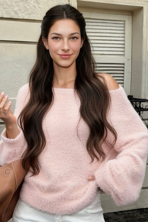 A woman wearing a fuzzy light pink off-the-shoulder sweater and white pants, holding a brown leather handbag and a lipstick, standing outdoors in front of a building wall.