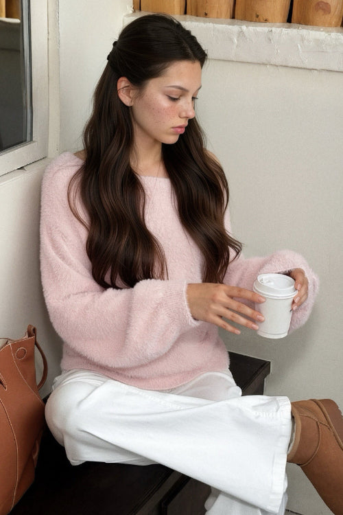 A woman with long dark hair sits on a dark wooden bench wearing a fuzzy light pink sweater and white wide-leg pants, holding a white paper coffee cup. A brown leather bag is on the bench next to her.