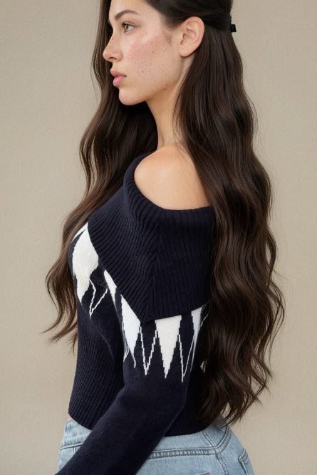 A woman wearing a dark navy off-the-shoulder knit sweater with a white geometric pattern on the front, paired with light blue jeans, shown from a side-back angle against a neutral beige background.