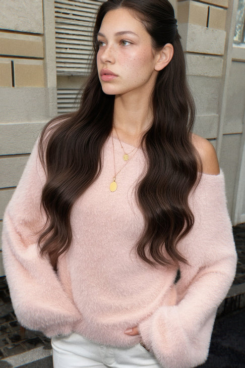 A woman with long, wavy dark brown hair wearing a fuzzy, pale pink off-the-shoulder sweater and white pants, standing outdoors against a light-colored stone wall.
