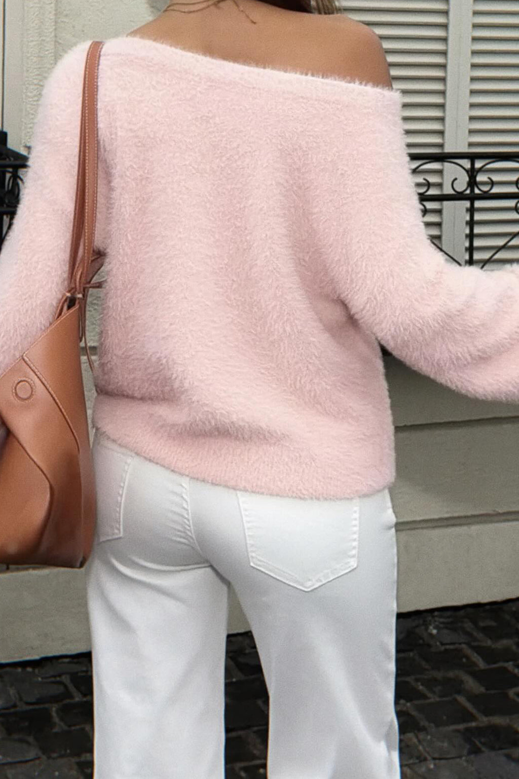 A woman wearing a fuzzy off-the-shoulder light pink sweater and white pants, carrying a brown leather shoulder bag, standing outdoors in front of a building with white shutters.