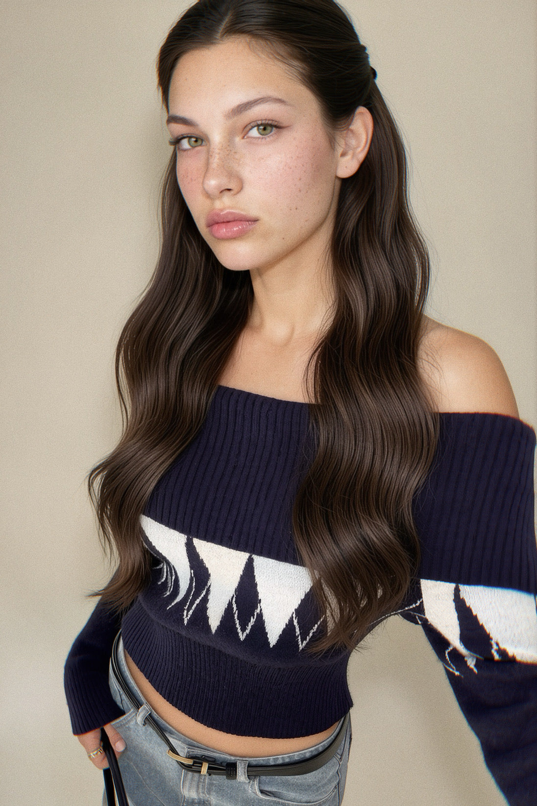 A woman modeling a navy blue off-the-shoulder knit sweater with a white geometric pattern across the chest and sleeves, paired with light-wash jeans against a plain beige background.