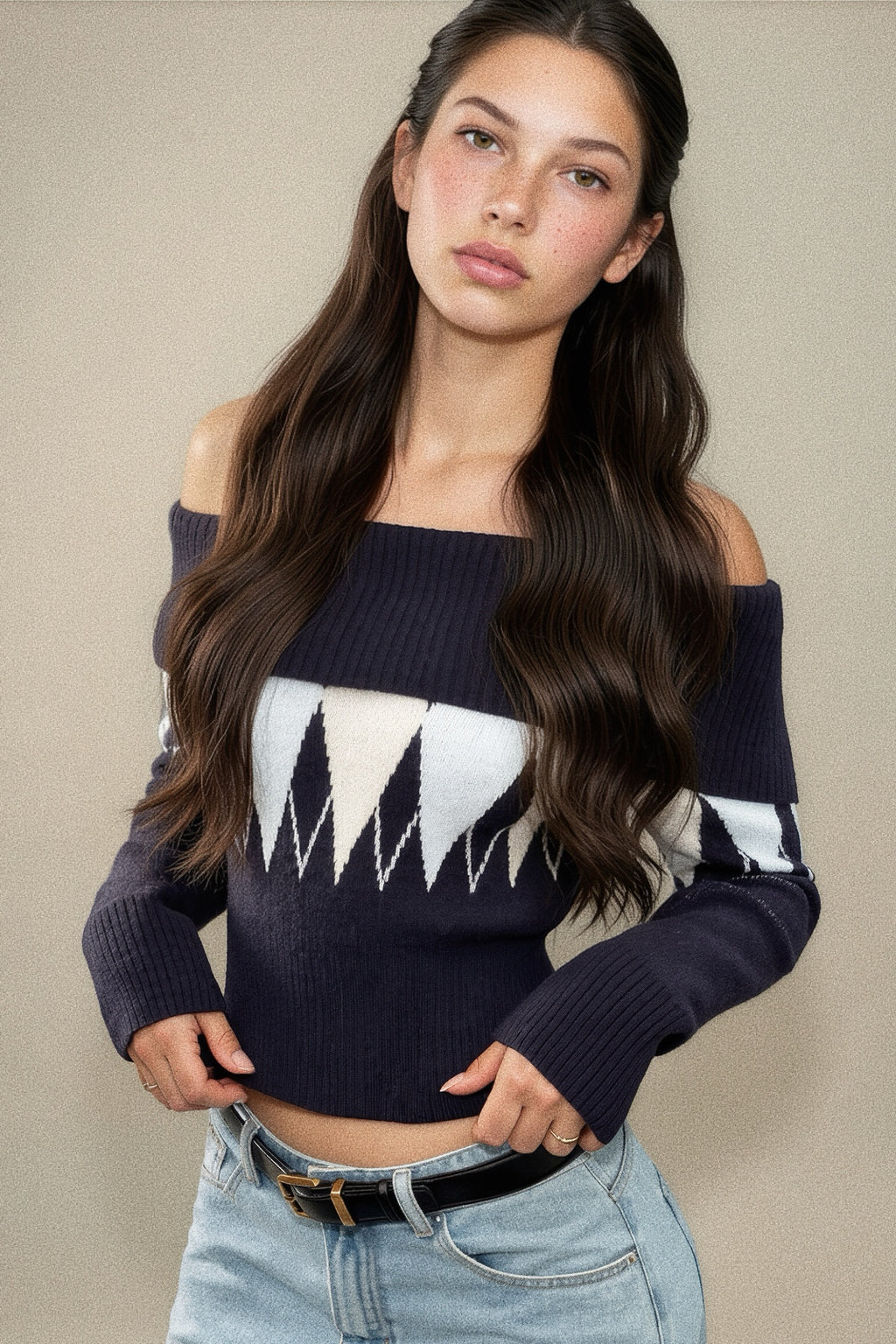 A woman modeling an off-the-shoulder navy blue knit sweater with a white geometric pattern across the chest and sleeves, paired with light-wash jeans against a neutral beige background.