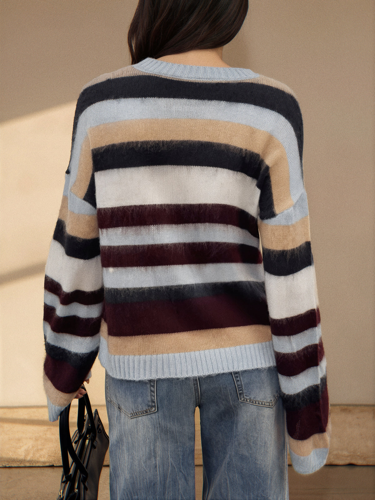 A person shown from the back wearing a fuzzy striped sweater with horizontal bands of cream, light blue, dark navy, burgundy, and tan, paired with blue jeans.