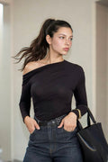 A woman wearing a black off-the-shoulder long-sleeve top and dark blue jeans, holding a black tote bag in a minimalist indoor setting.