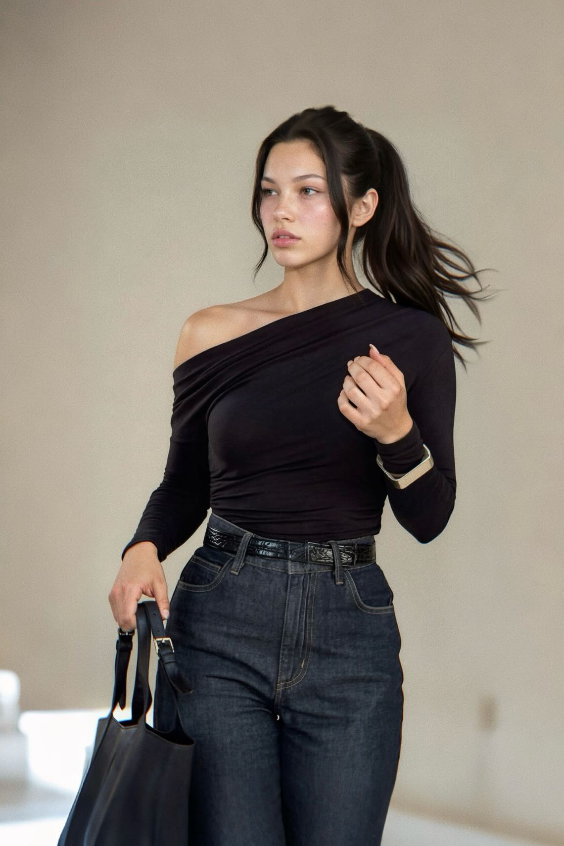 A woman wearing a black off-the-shoulder long-sleeve top and dark blue jeans, holding a black tote bag against a neutral beige background.