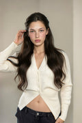 A woman with long dark hair wearing a cream-colored asymmetrical cardigan and dark jeans, posing in front of a neutral beige wall with a window on the left.