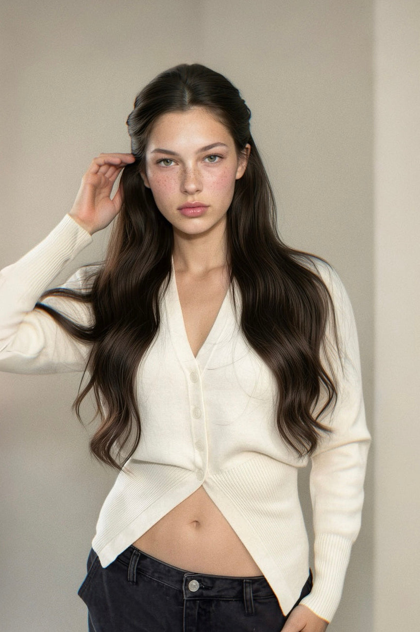 A woman with long dark hair wearing a cream-colored asymmetrical cardigan and dark jeans, posing in front of a neutral beige wall with a window on the left.