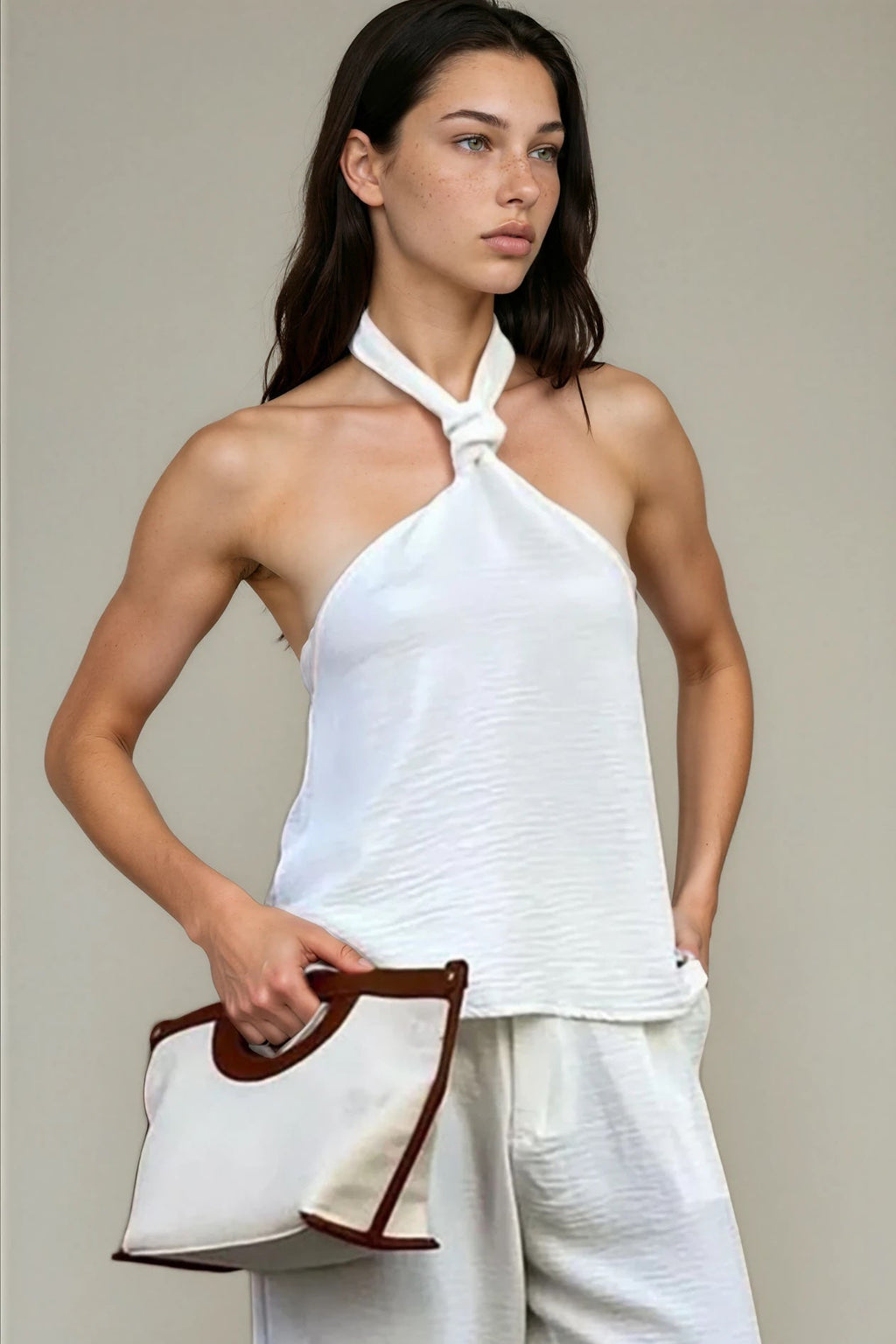 Woman wearing a white halter top and pants, holding a matching bag against a neutral background