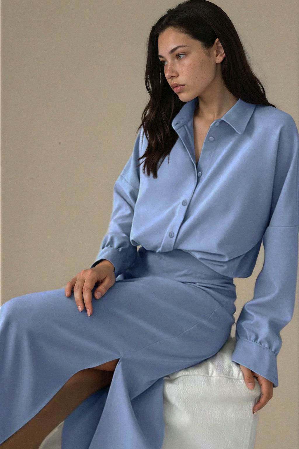Woman wearing a light blue shirt and pants sitting on a white stool against a beige wall.