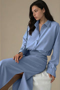 Woman wearing a light blue shirt and pants sitting on a white stool against a beige wall.