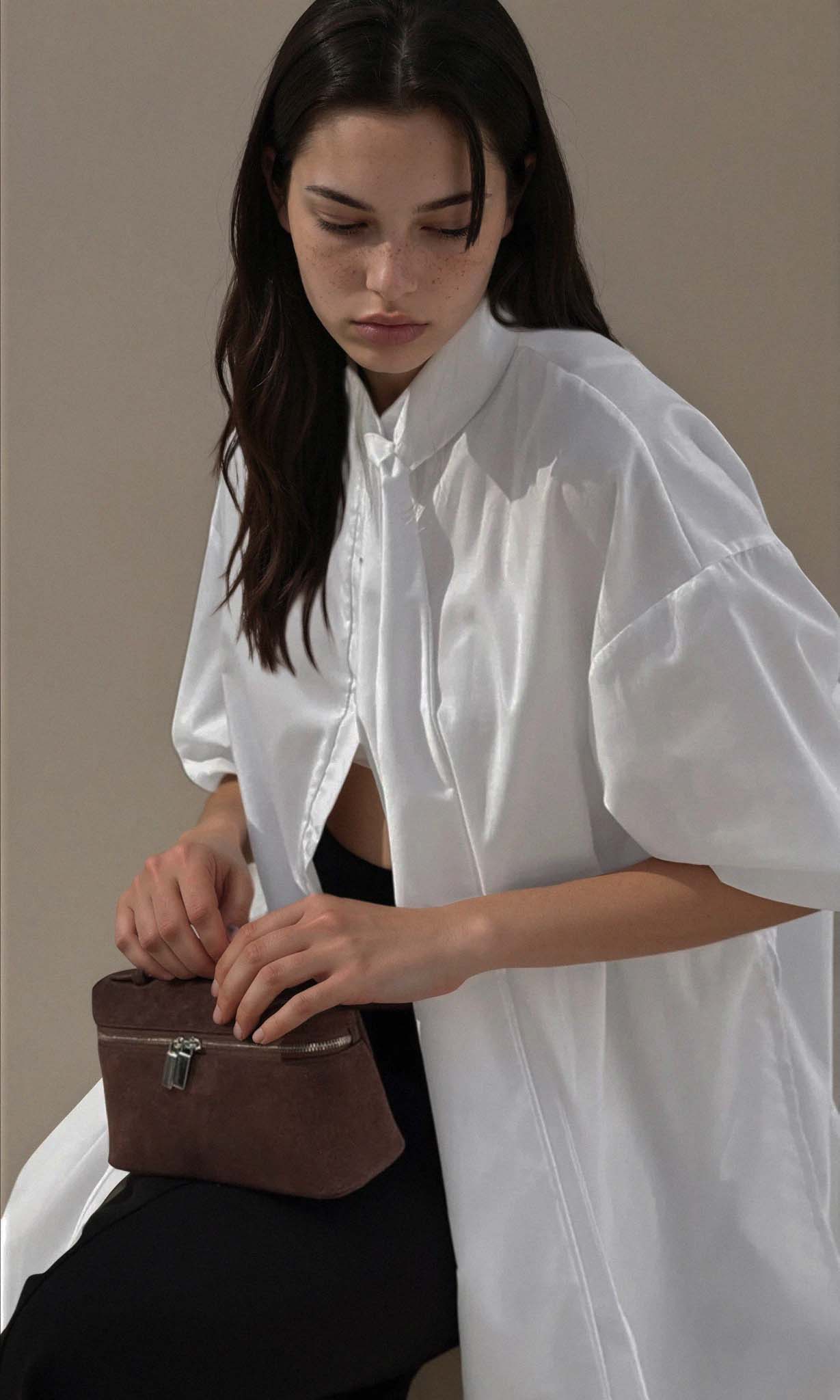 Woman holding a brown leather bag wearing a white shirt against a neutral background