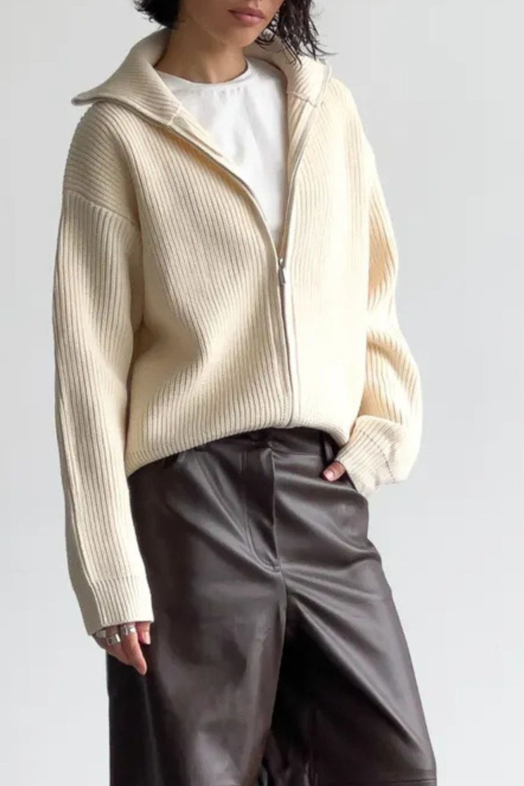 Person wearing a cream ribbed sweater over a white shirt with dark pants on a light gray background