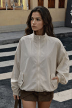 A woman wearing a light beige zip-up jacket with a stand-up collar and front pockets, paired with brown shorts, standing on a street crosswalk.