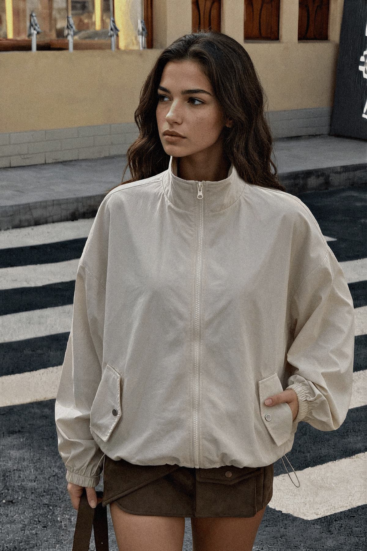 A woman wearing a light beige zip-up jacket with a stand-up collar and front pockets, paired with brown shorts, standing on a street crosswalk.
