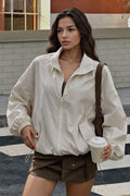 A woman wearing an oversized off-white zip-up jacket and brown shorts, holding a takeaway coffee cup and wearing a brown shoulder bag, standing outdoors on a street.