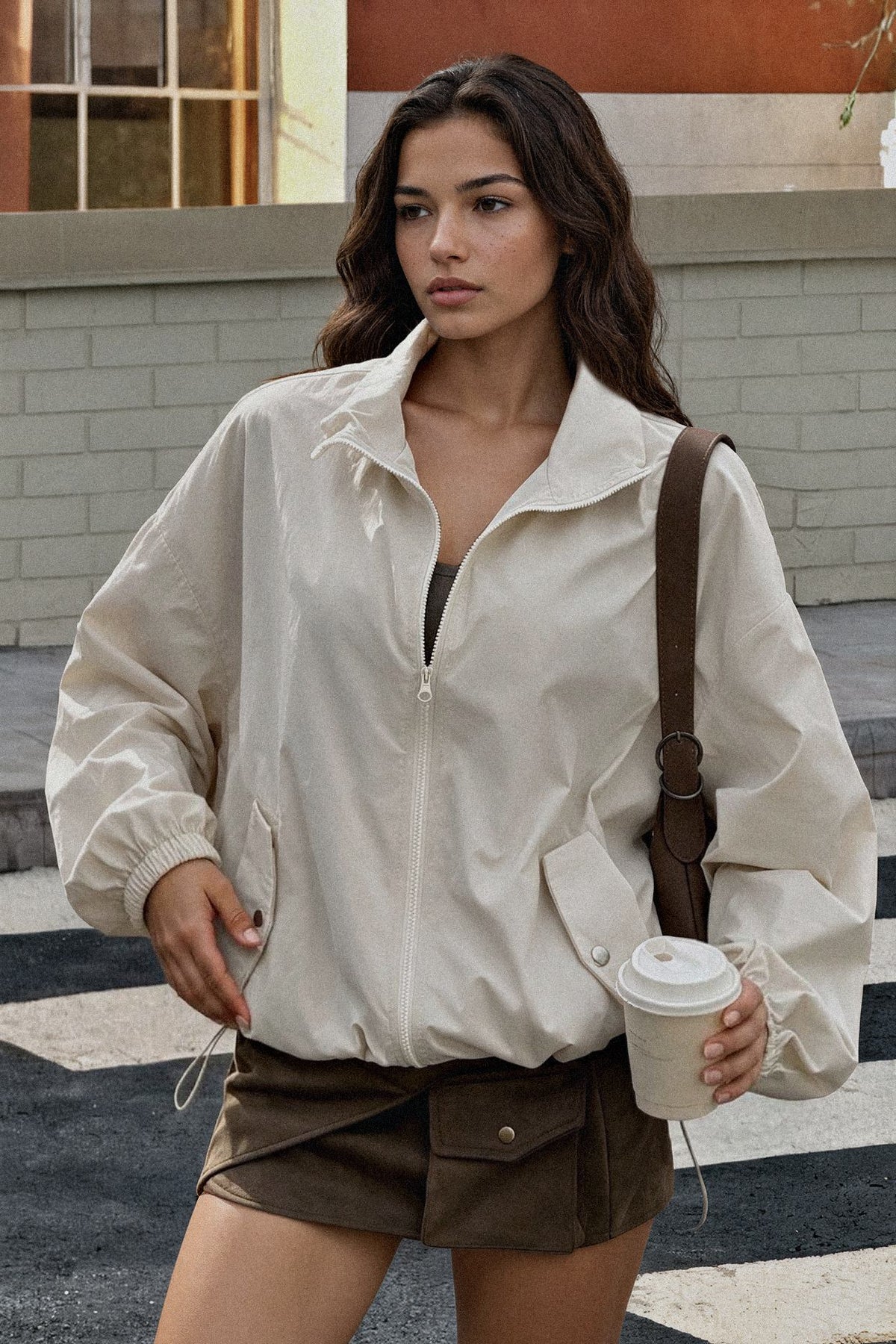 A woman wearing an oversized off-white zip-up jacket and brown shorts, holding a takeaway coffee cup and wearing a brown shoulder bag, standing outdoors on a street.