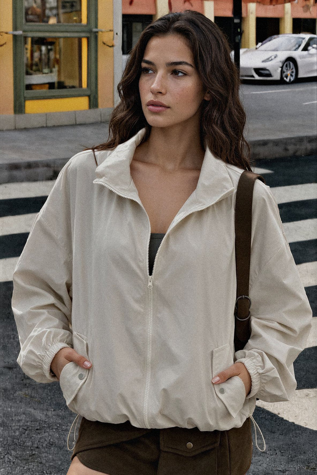 A woman wearing a light beige zip-up windbreaker jacket with elastic cuffs and a stand collar, standing on a city street crosswalk.