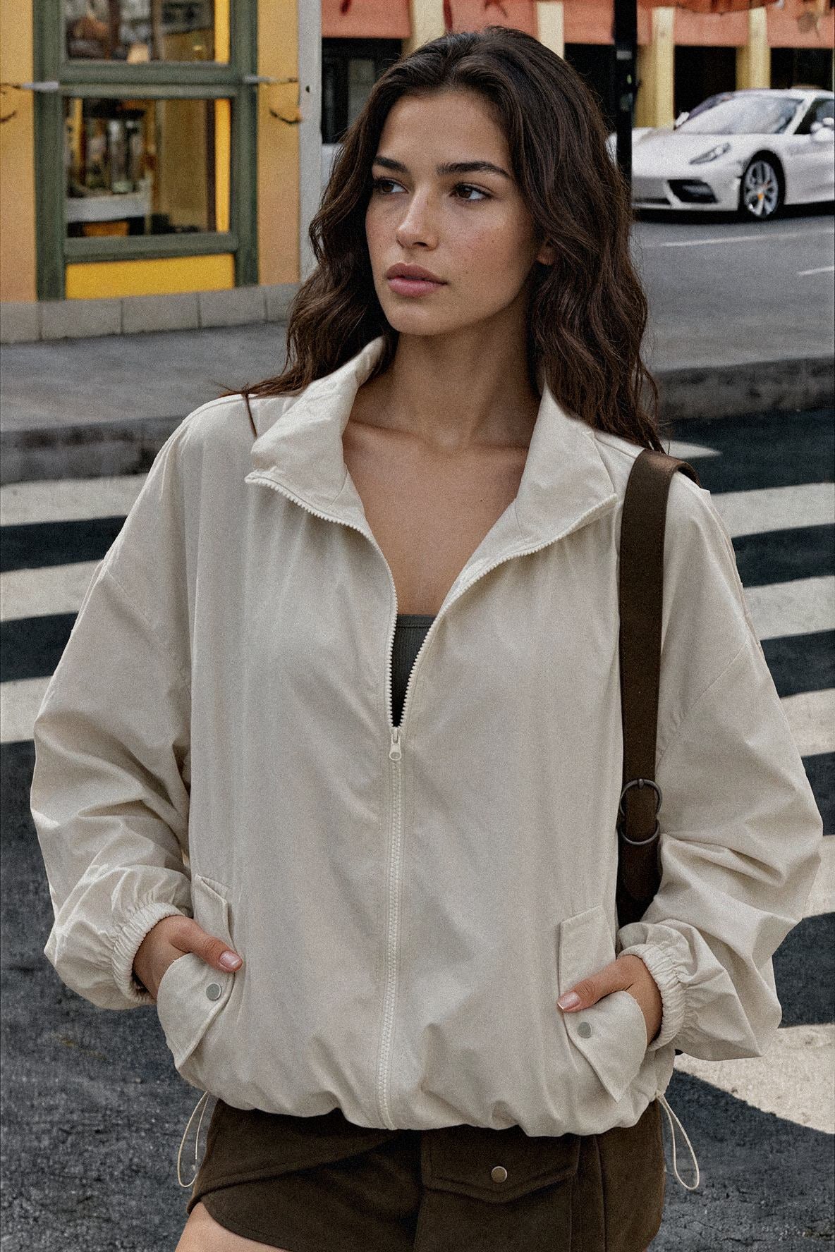 A woman wearing a light beige zip-up windbreaker jacket with elastic cuffs and a stand collar, standing on a city street crosswalk.