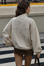 A woman viewed from behind wearing an oversized cream-colored lightweight jacket and a brown mini skirt, standing on a city street.