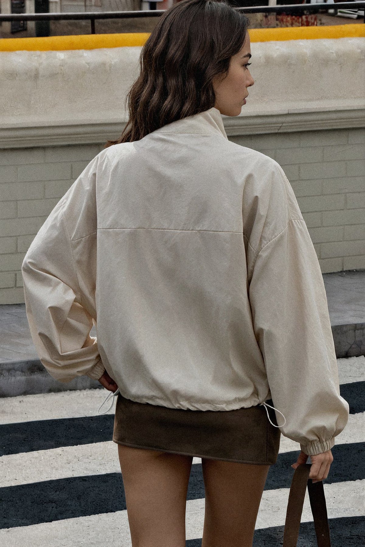 A woman viewed from behind wearing an oversized cream-colored lightweight jacket and a brown mini skirt, standing on a city street.