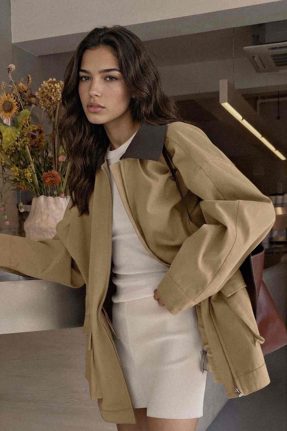 A woman wearing an oversized tan utility jacket with dark contrast shoulder panels over a white ribbed mini dress, standing in a modern indoor setting.