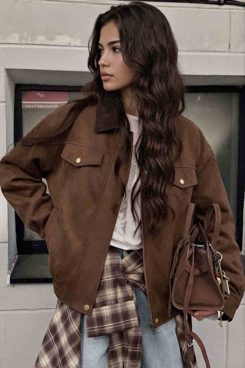 A woman wearing an oversized brown suede jacket over a white top and plaid shirt, standing outdoors near a building wall and holding a brown handbag.