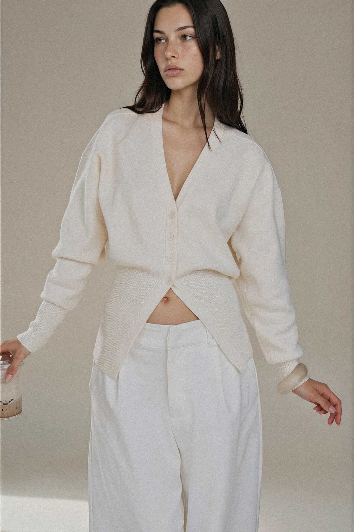 A woman modeling a cream-colored knit cardigan with a deep V-neck and button front, paired with matching cream wide-leg trousers, against a neutral beige studio background.
