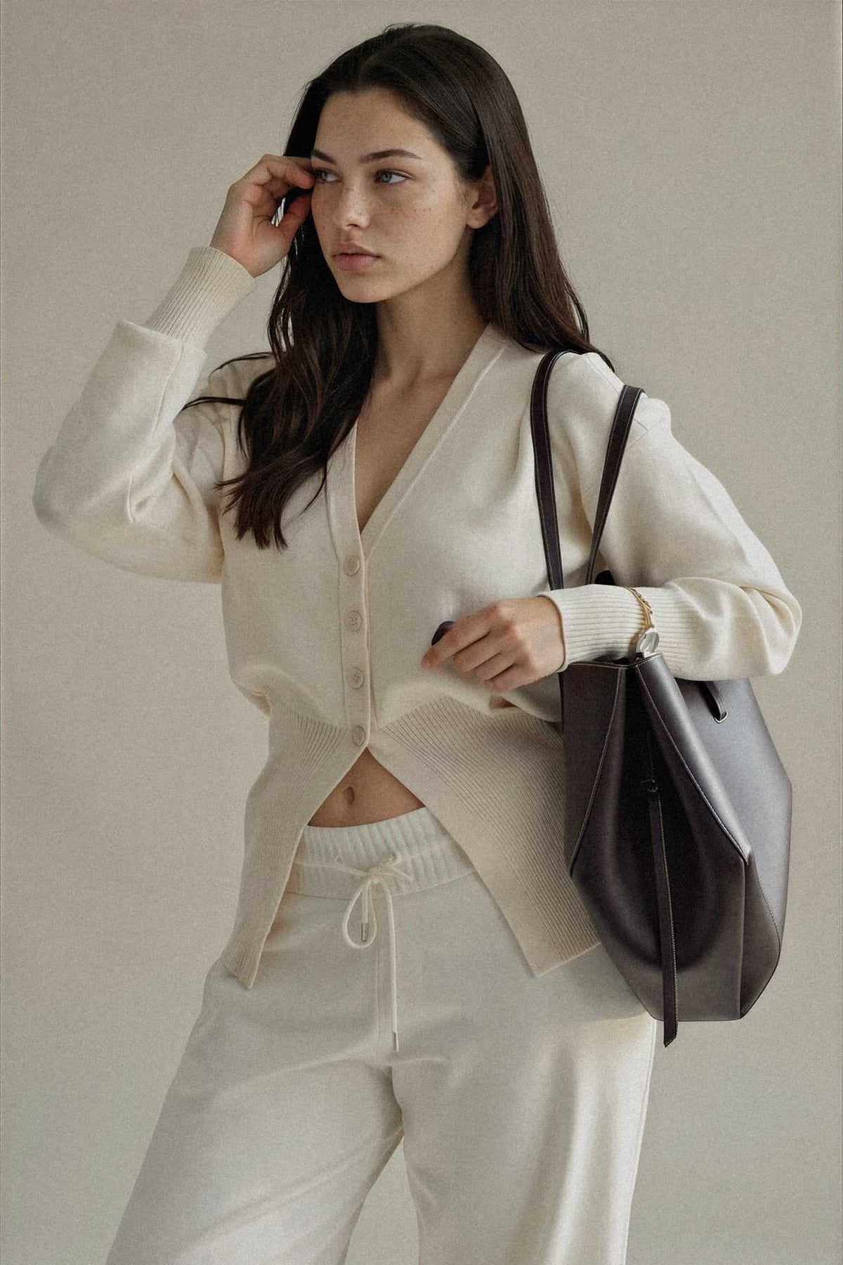 A woman modeling a cream-colored knit cardigan and matching sweatpants, carrying a dark brown leather shoulder bag against a neutral beige background.