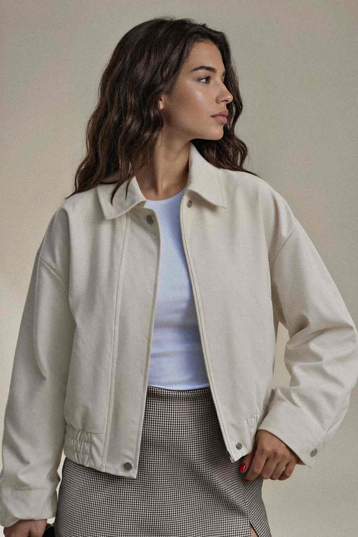 A woman wearing an oversized off-white cropped jacket over a light blue top and a gray houndstooth skirt, photographed against a plain beige studio background.