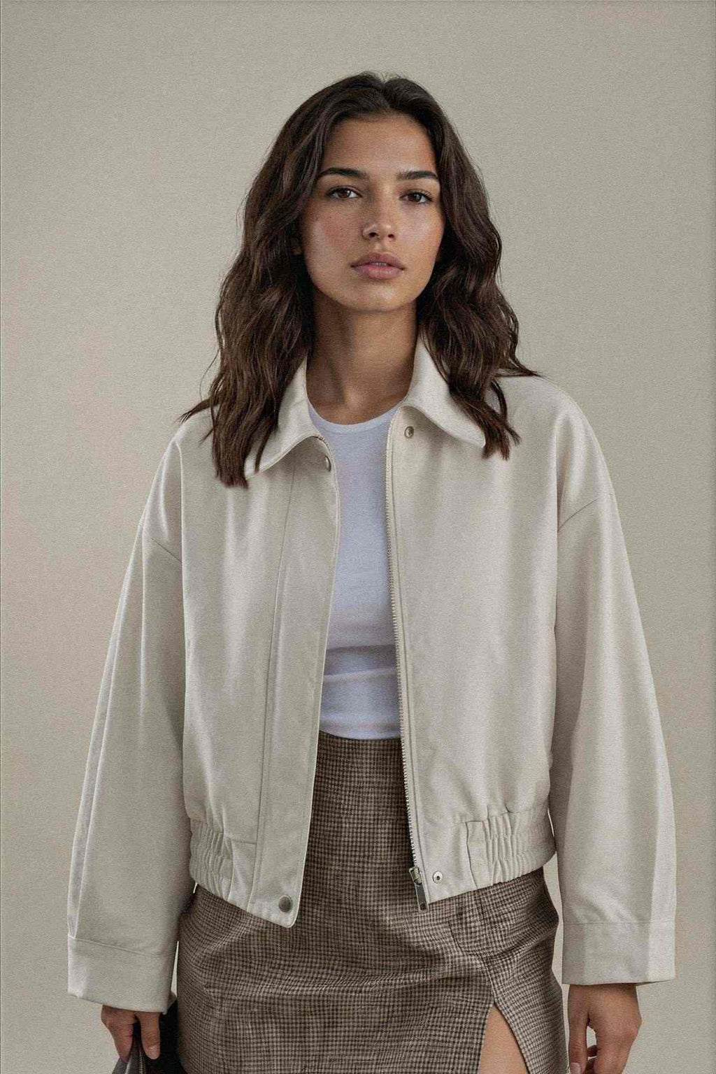 A woman modeling a light beige cropped jacket over a white top and brown plaid skirt, set against a plain beige studio background.