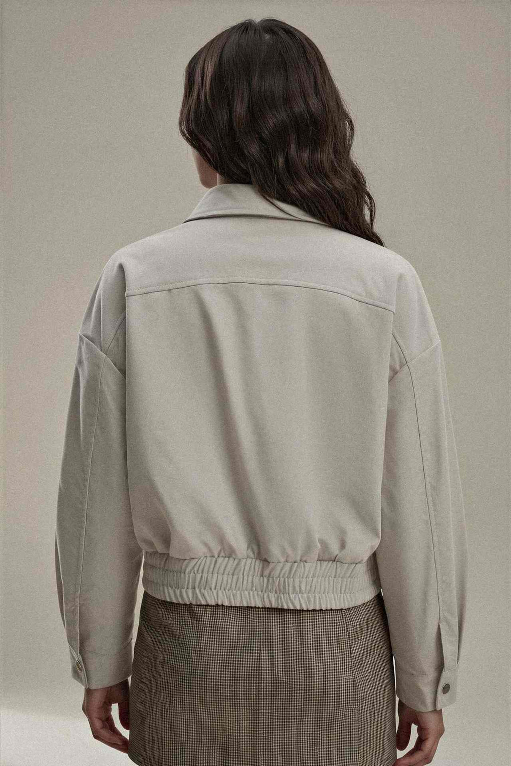A model shown from the back wearing a light beige cropped jacket with a gathered elastic waist, paired with a brown plaid skirt, against a neutral beige background.