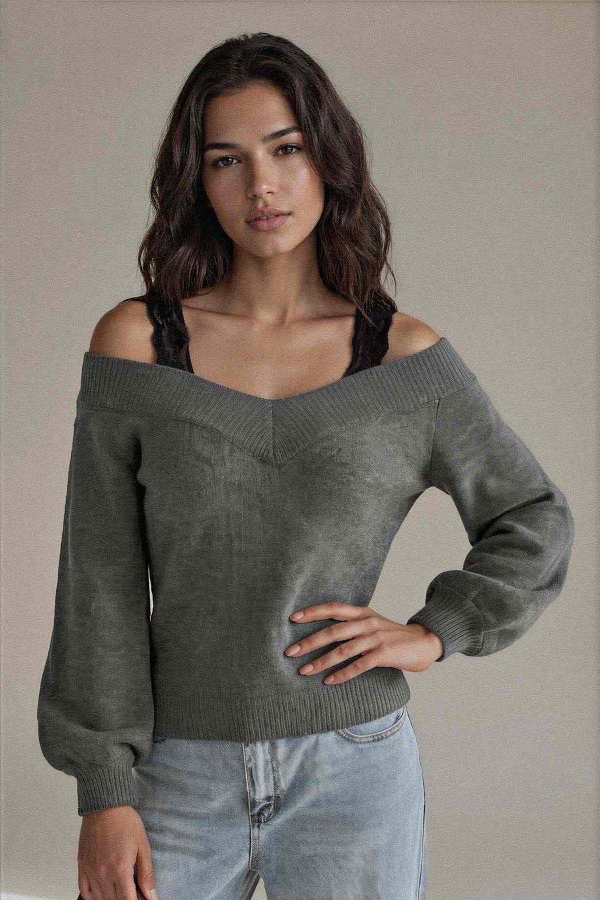 A woman modeling an off-the-shoulder knit sweater in a muted gray-green color, paired with light-wash jeans, against a plain neutral background.