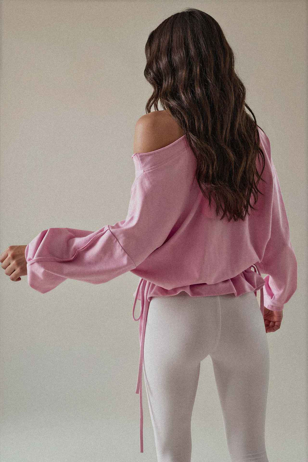 A woman shown from the back wearing a light pink off-the-shoulder long-sleeve top with a drawstring waist and white leggings, set against a plain light beige studio background.