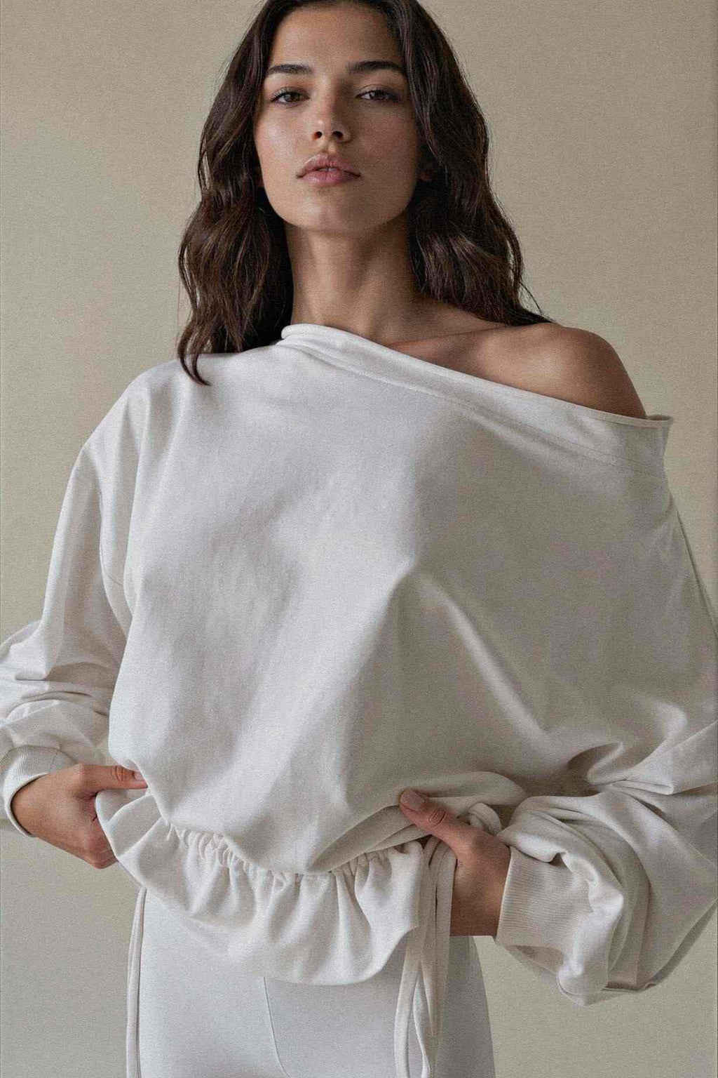 A woman wearing a white off-the-shoulder long-sleeve top with a gathered ruffle hem, photographed against a plain beige studio background.