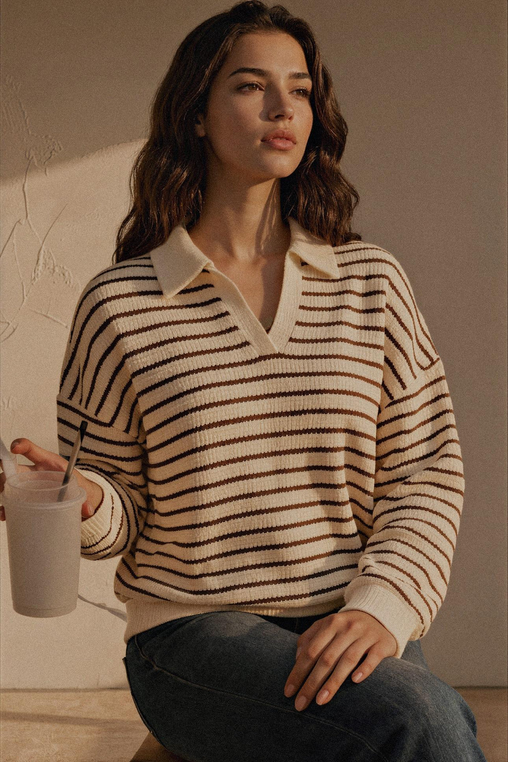 A woman wearing a cream and brown horizontally striped knit polo sweater with a V-neck collar, paired with dark blue jeans, holding a drink against a warm beige wall.