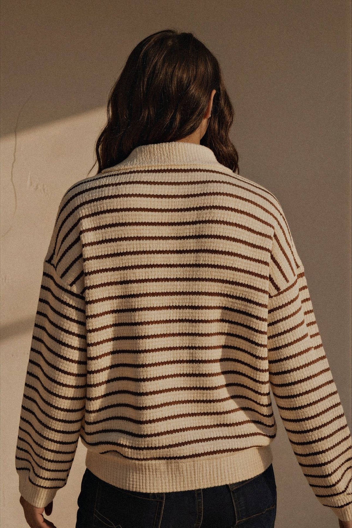 A person viewed from behind wearing a cream knit sweater with thin dark brown horizontal stripes and dark blue jeans, standing against a plain beige wall in warm directional light.