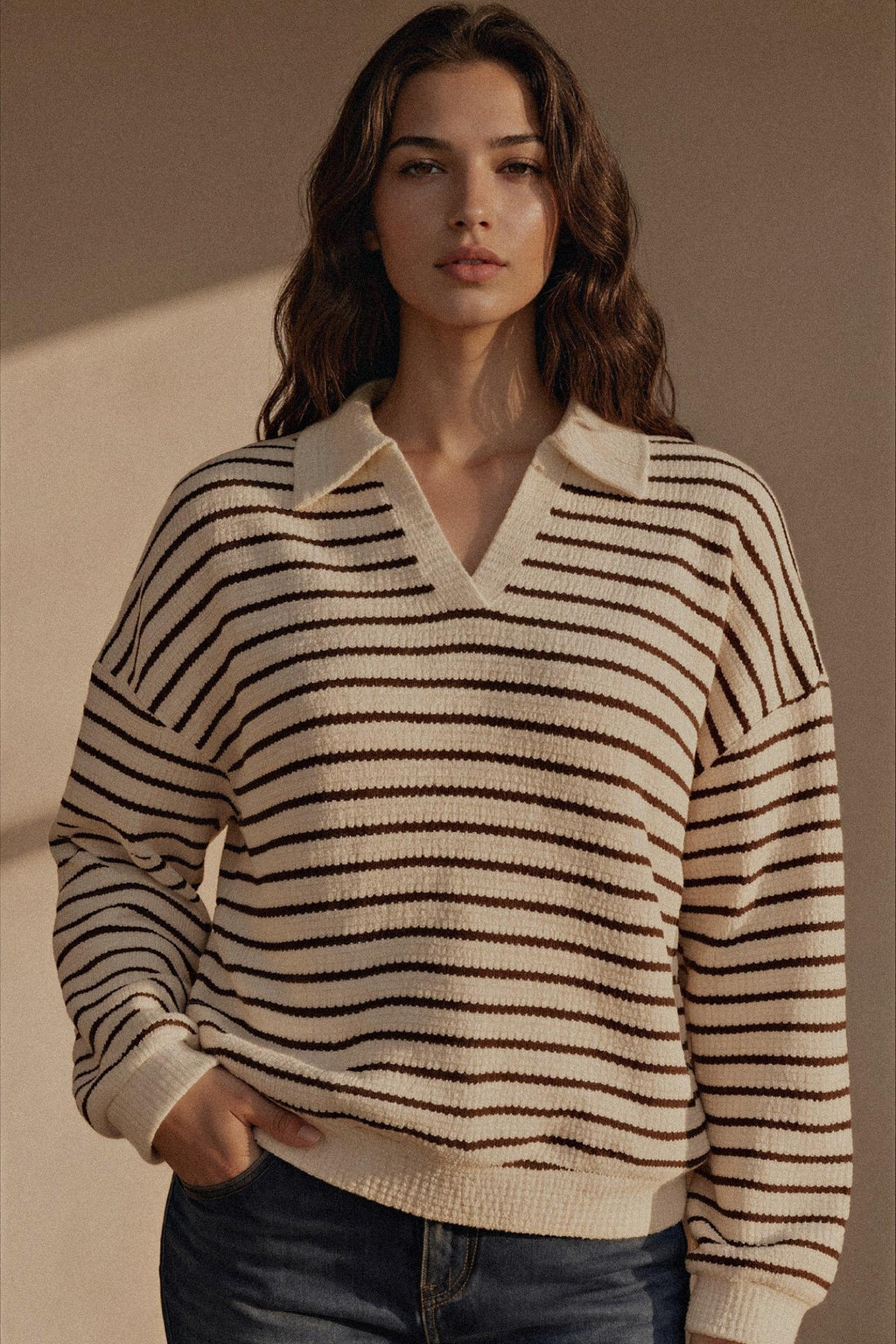 A woman wearing a cream and dark brown horizontally striped knit sweater with a V-neck collar, paired with dark blue jeans, set against a warm beige background with soft shadows.