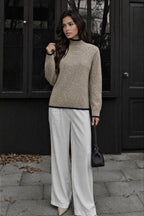 A woman stands outdoors in front of a dark building, wearing a beige knit mock-neck sweater with dark trim, white wide-leg pants, and holding a small black handbag.